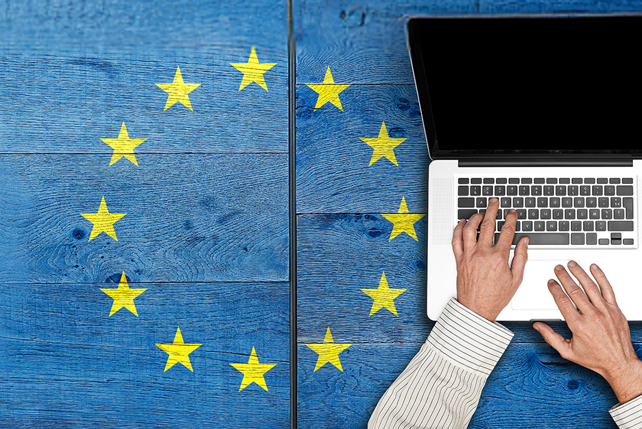 Person tippt auf einer Laptop-Tastatur auf einem blauen Holztisch, umgeben von gelben Sternen der EU Flagge.