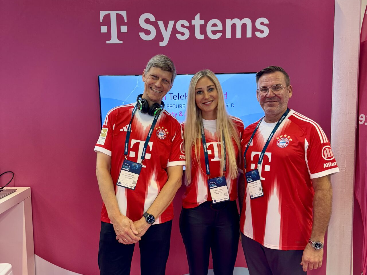 Drei Personen in FC Bayern München Trikots posieren am T-Systems Stand vor einem Bildschirm
