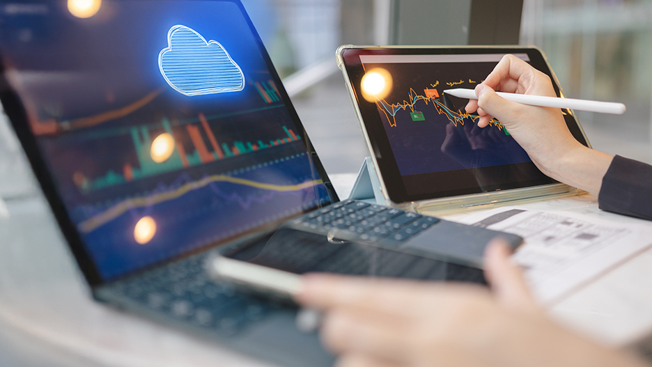  Zwei geöffnete Laptops: Im Vordergrund hält eine Hand ein Smartphone, im Hintergrund arbeitet jemand mit einem Stift am Laptop.