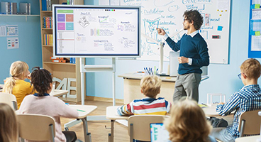 Pupils sit in a classroom, a teacher stands in front of a digital whiteboard 